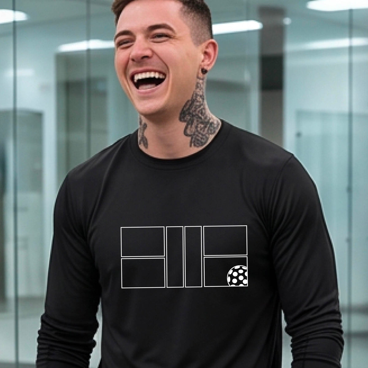 A smiling man wearing a black long-sleeve athletic shirt with a white geometric pickleball court and ball graphic on the chest,.