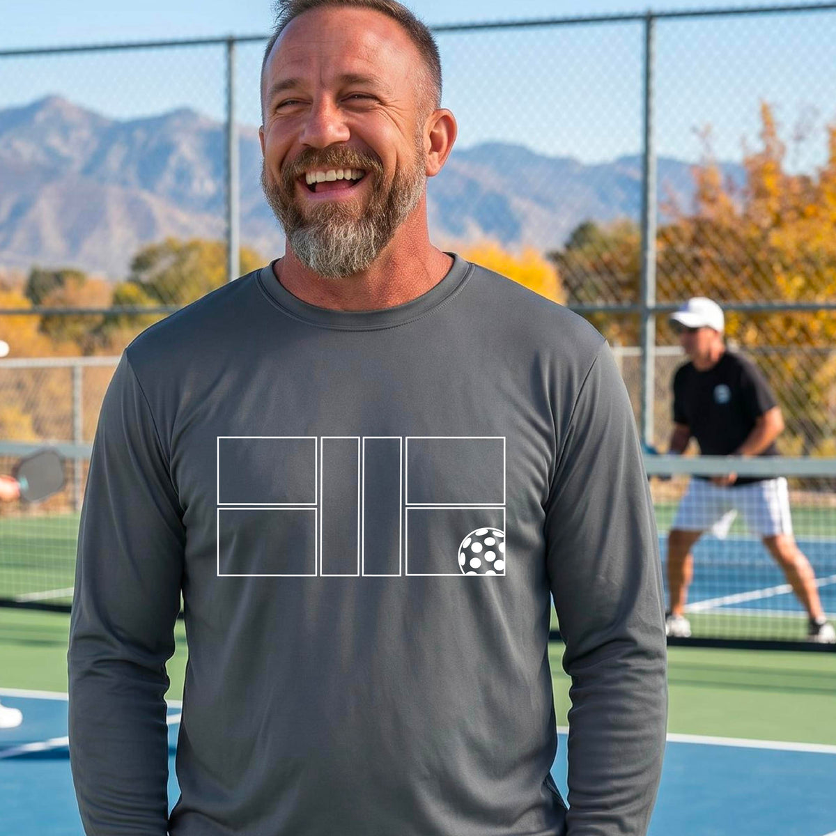 A smiling man wearing a gray long-sleeve athletic shirt with a white geometric pickleball court and ball graphic on the chest,.