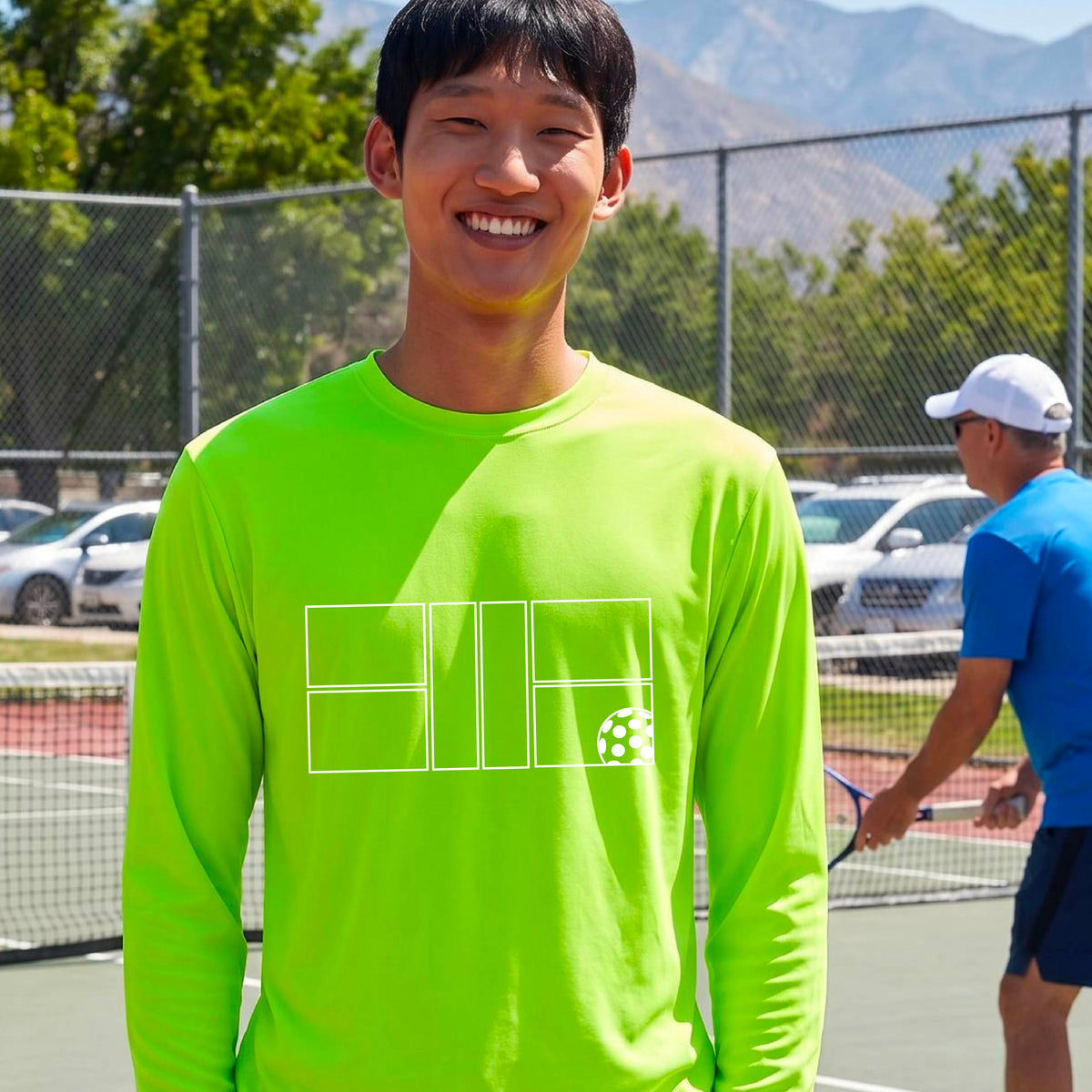 A smiling man wearing a lime green long-sleeve athletic shirt with a white geometric pickleball court and ball graphic on the chest,.
