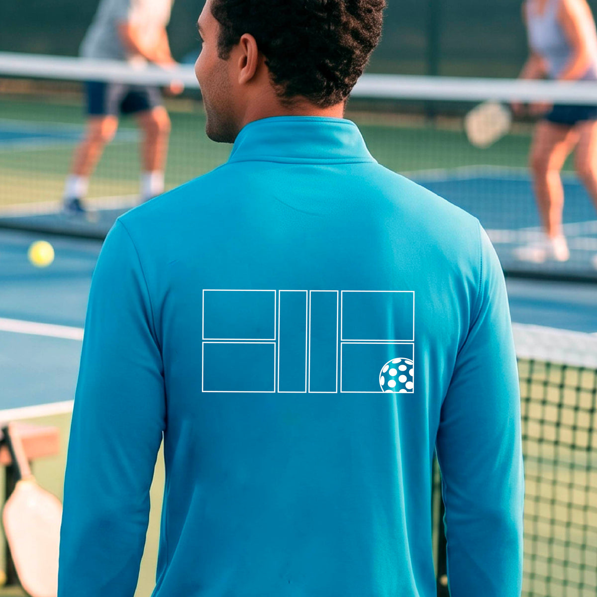 A man wearing a bright blue quarter-zip shirt on a pickleball court with a white graphic of a pickleball court and ball graphic on the back of the shirt, standing on a pickleball court. 