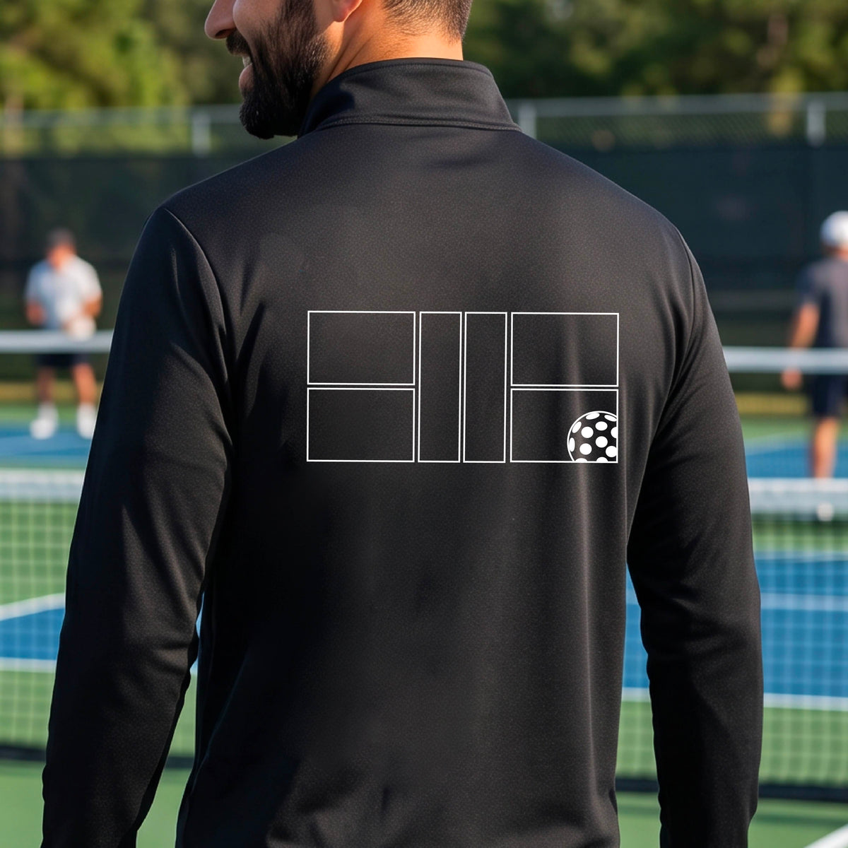 A man wearing a black quarter-zip shirt on a pickleball court with a white graphic of a pickleball court and ball graphic on the back of the shirt, standing on a pickleball court. 