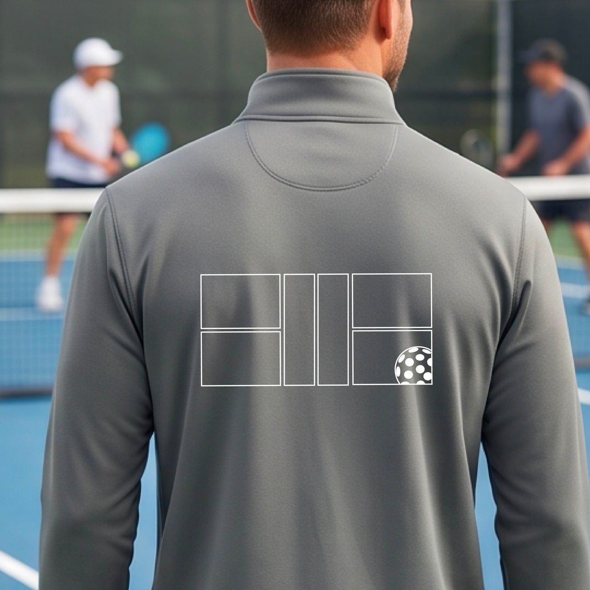 A man wearing a gray quarter-zip shirt on a pickleball court with a white graphic of a pickleball court and ball graphic on the back of the shirt, standing on a pickleball court. 