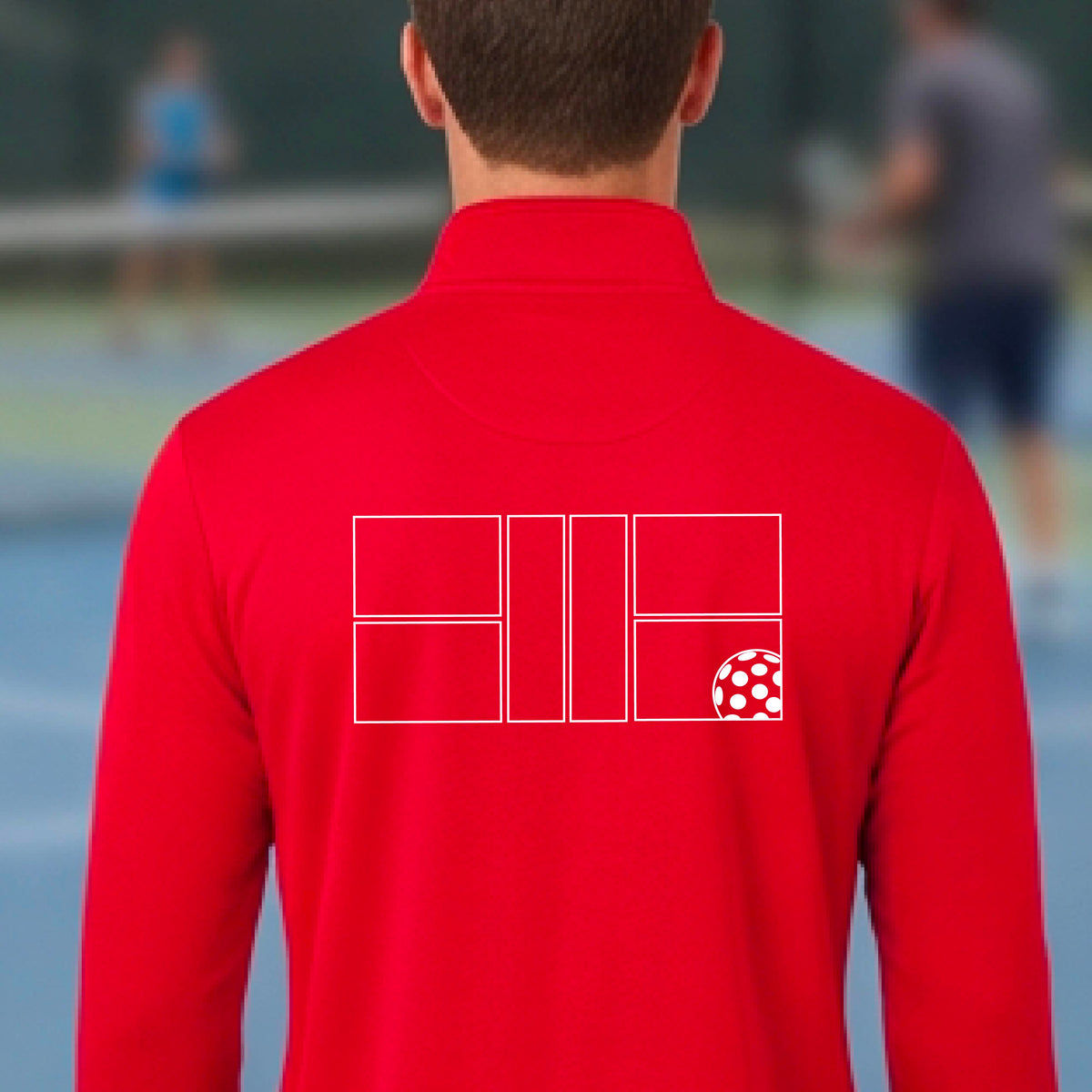 A man wearing a red quarter-zip shirt on a pickleball court with a white graphic of a pickleball court and ball graphic on the back of the shirt, standing on a pickleball court. 