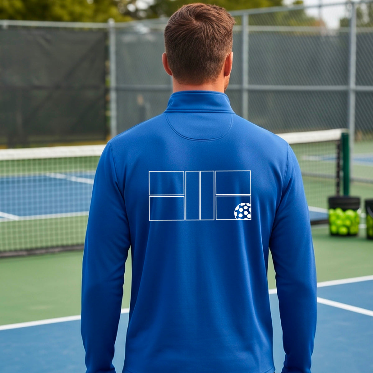 A man wearing a royal blue quarter-zip shirt on a pickleball court with a white graphic of a pickleball court and ball graphic on the back of the shirt, standing on a pickleball court. 