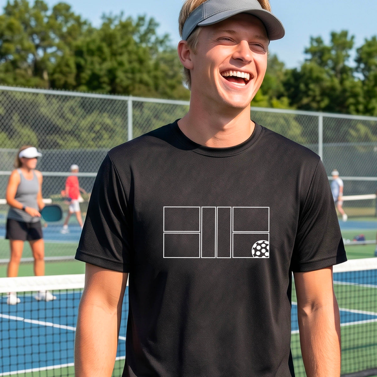 A man wearing a black short-sleeve shirt with a graphic of a white pickleball court and ball on the front, standing on a pickleball court. 