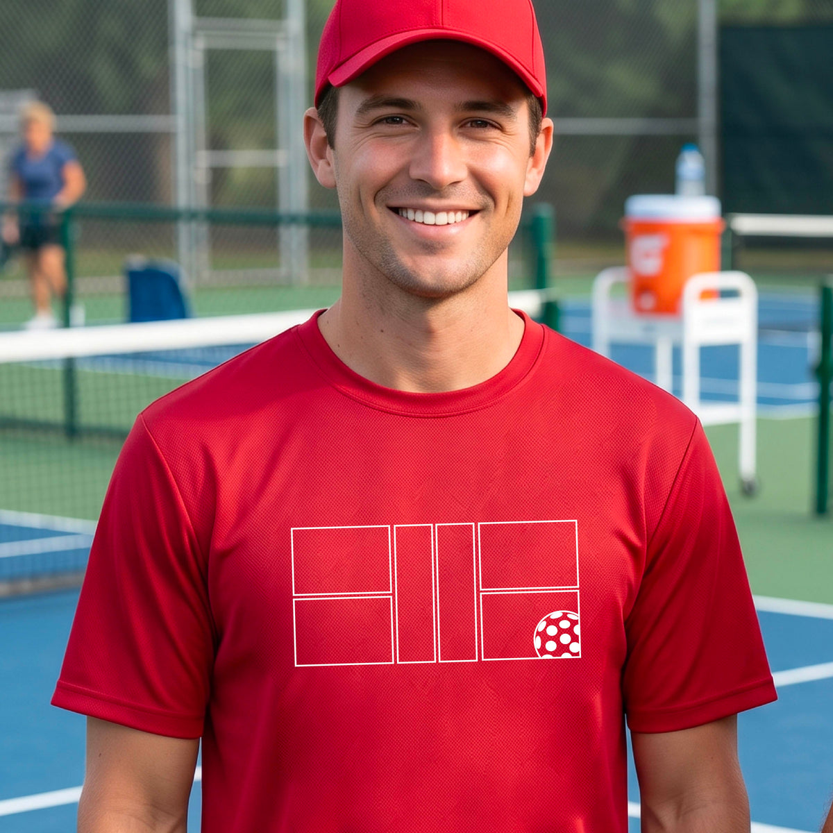 A man wearing a redl short-sleeve shirt with a graphic of a white pickleball court and ball on the front, standing on a pickleball court. 