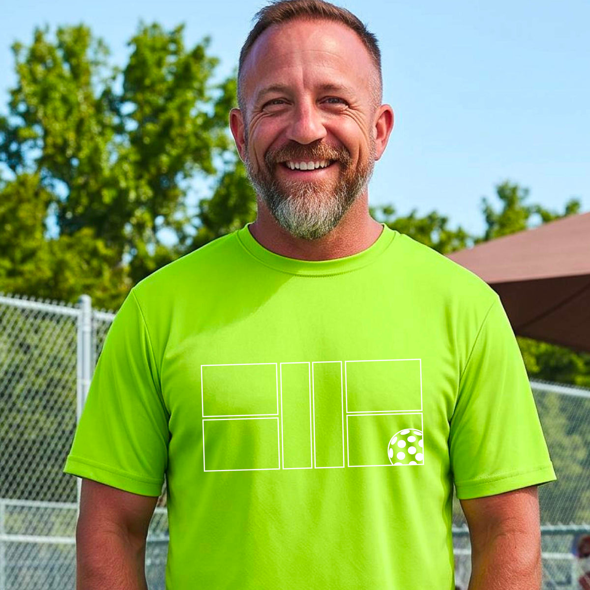 A man wearing a lime green short-sleeve shirt with a graphic of a white pickleball court and ball on the front, standing on a pickleball court. 