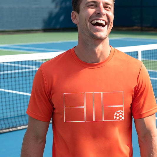 A man wearing an orange short-sleeve shirt with a graphic of a white pickleball court and ball on the front, standing on a pickleball court. 
