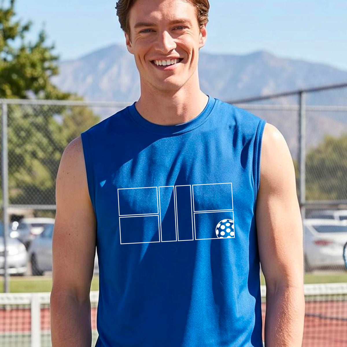  A man wearing a royal blue sleeveless shirt with a white pickleball court and ball graphic design, standing on a pickleball court. 