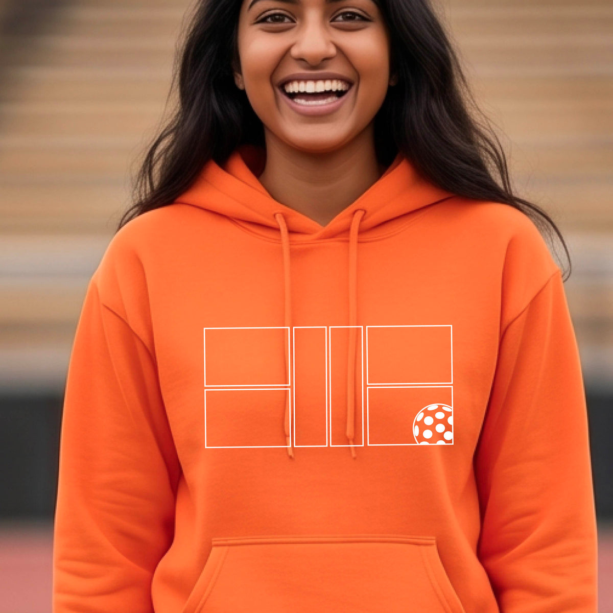 A man wearing an orange unisex hoodie pullover with a white pickleball court and all graphic on the front of the sweatshirt, standing on a pickleball court. 