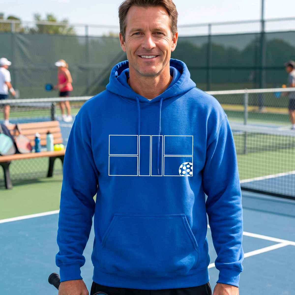 A man wearing a royal blue unisex hoodie pullover with a white pickleball court and all graphic on the front of the sweatshirt, standing on a pickleball court. 