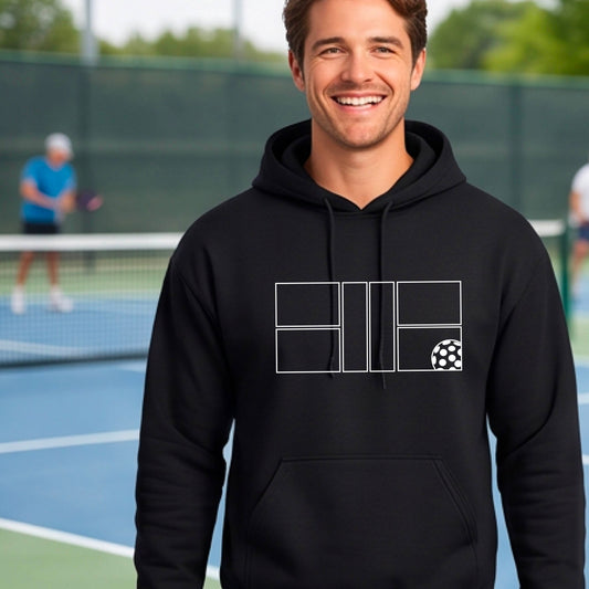A man wearing a black unisex hoodie pullover with a white pickleball court and all graphic on the front of the sweatshirt, standing on a pickleball court. 
