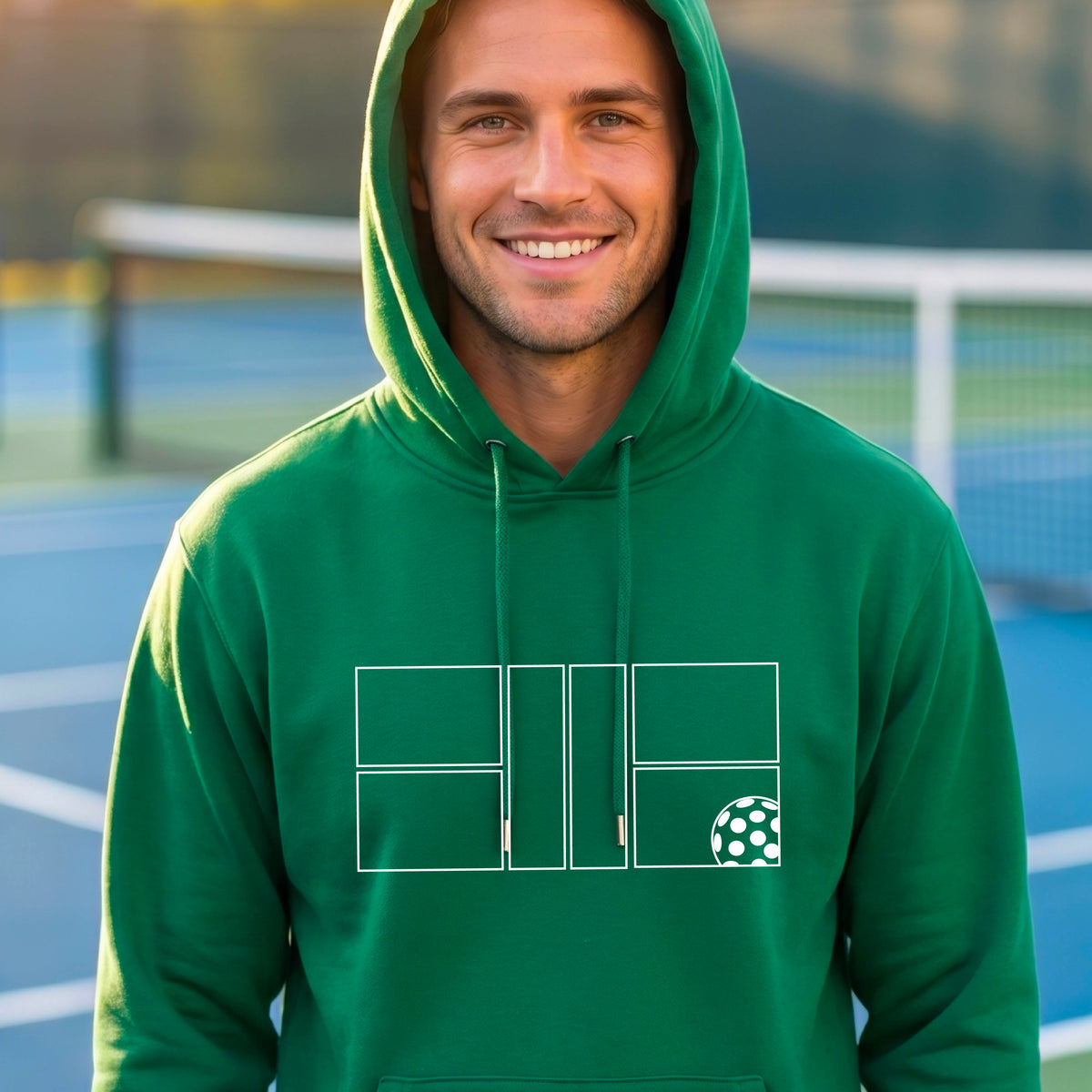A man wearing a green unisex hoodie pullover with a white pickleball court and all graphic on the front of the sweatshirt, standing on a pickleball court. 