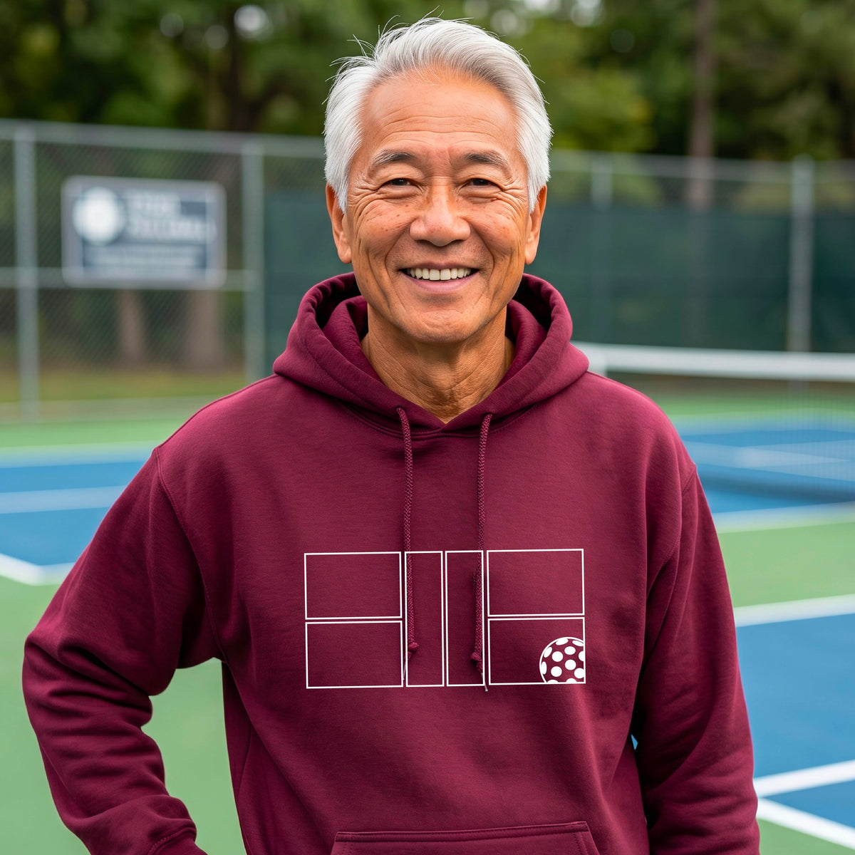 A man wearing a maroon unisex hoodie pullover with a white pickleball court and all graphic on the front of the sweatshirt, standing on a pickleball court. 