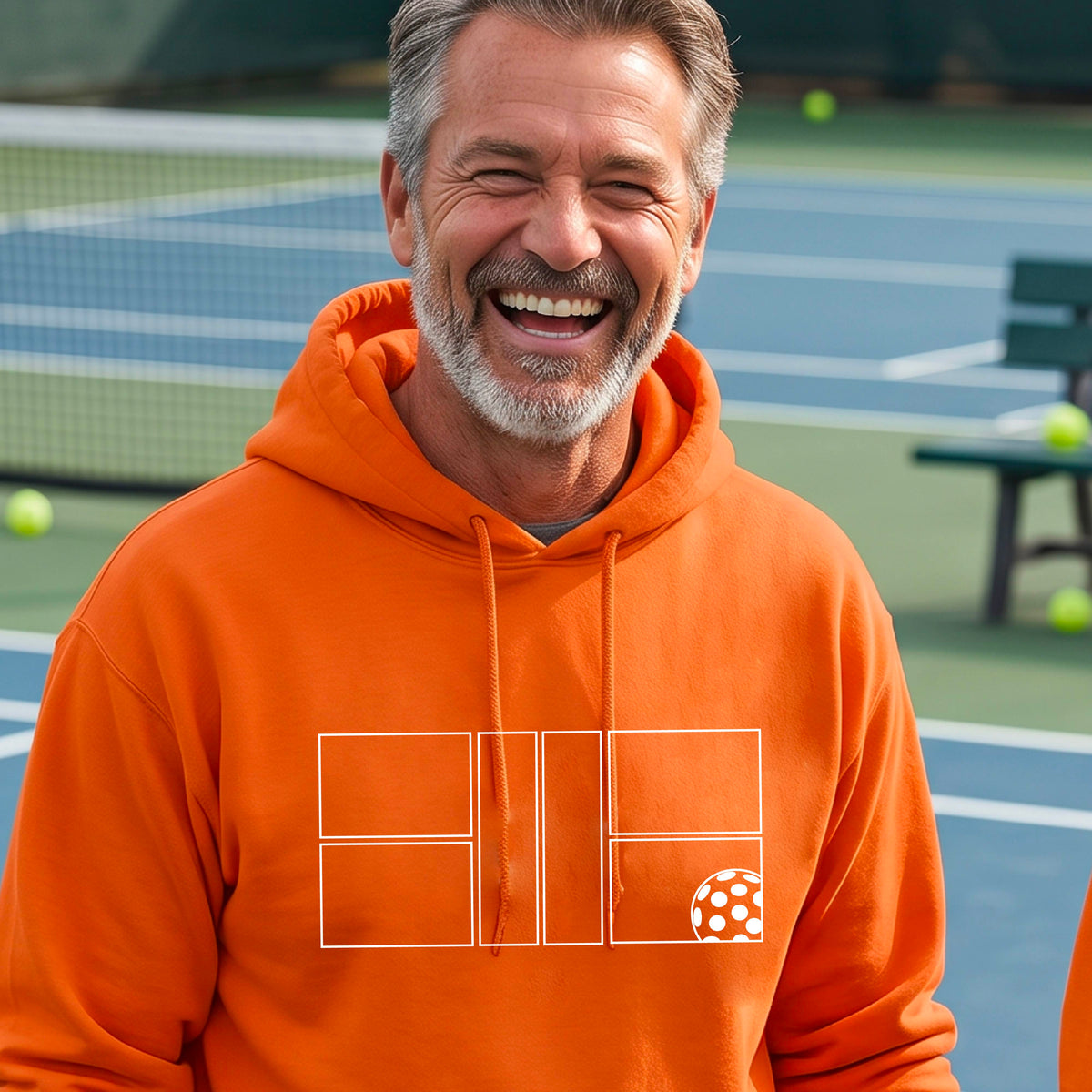 A man wearing an orange unisex hoodie pullover with a white pickleball court and all graphic on the front of the sweatshirt, standing on a pickleball court. 