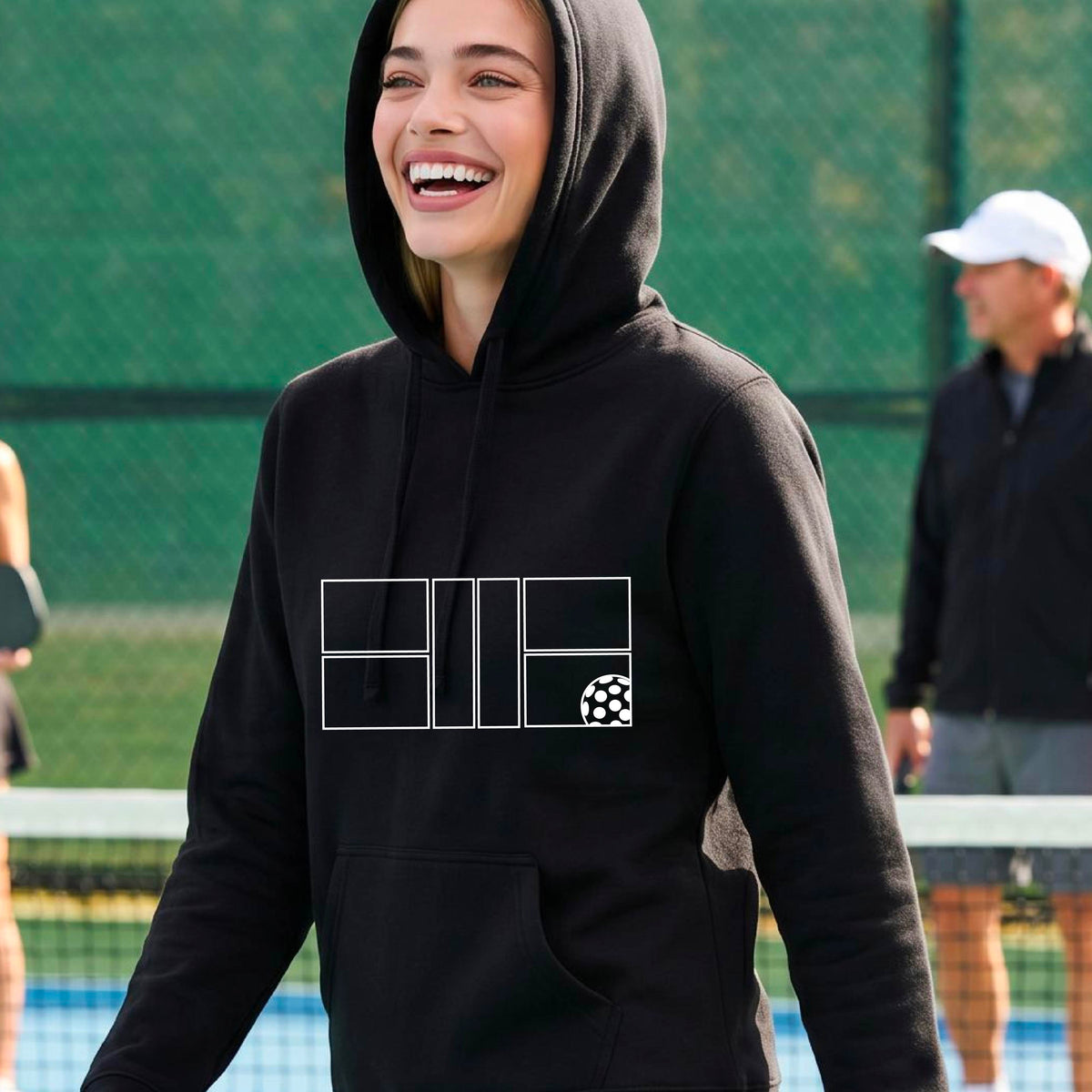 A woman wearing a black pullover hoodie with a white geometric pickleball court line-art graphic and a small pickleball ball icon on the chest.