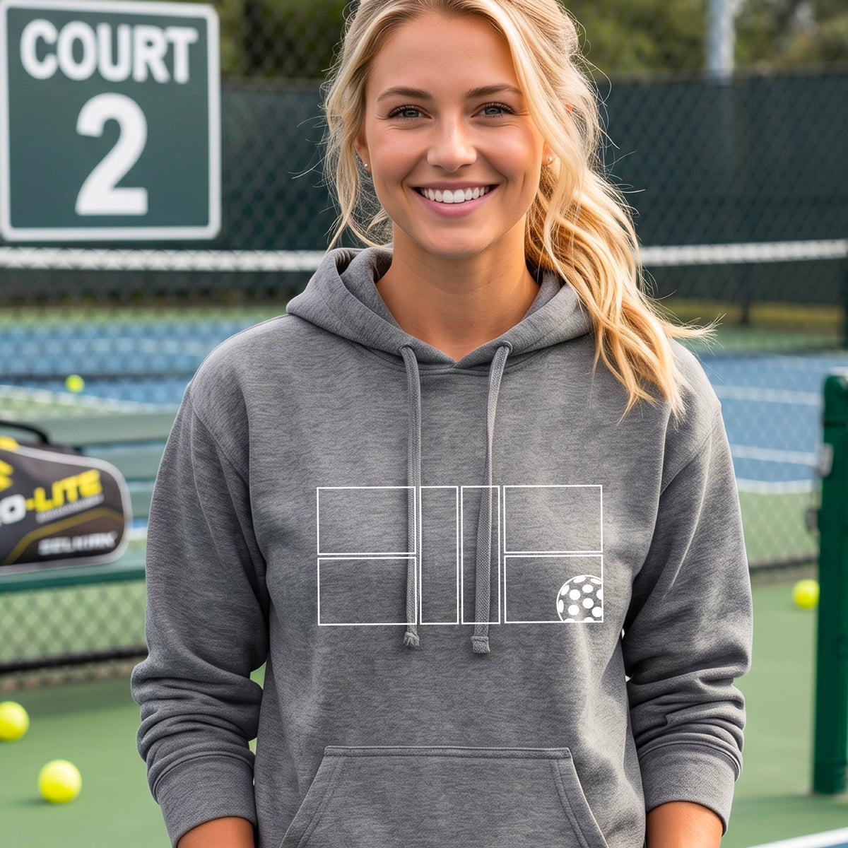A woman wearing a gray pullover hoodie with a white geometric pickleball court line-art graphic and a small pickleball ball icon on the chest.