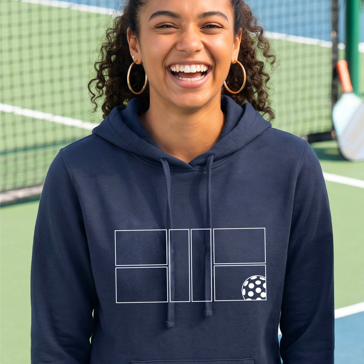 A woman wearing a navy blue pullover hoodie with a white geometric pickleball court line-art graphic and a small pickleball ball icon on the chest.