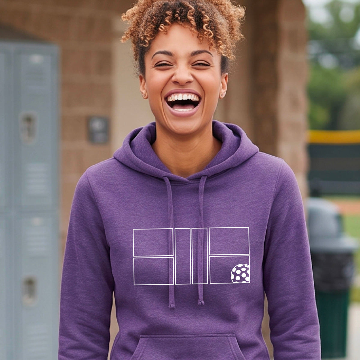A woman wearing a purple pullover hoodie with a white geometric pickleball court line-art graphic and a small pickleball ball icon on the chest.