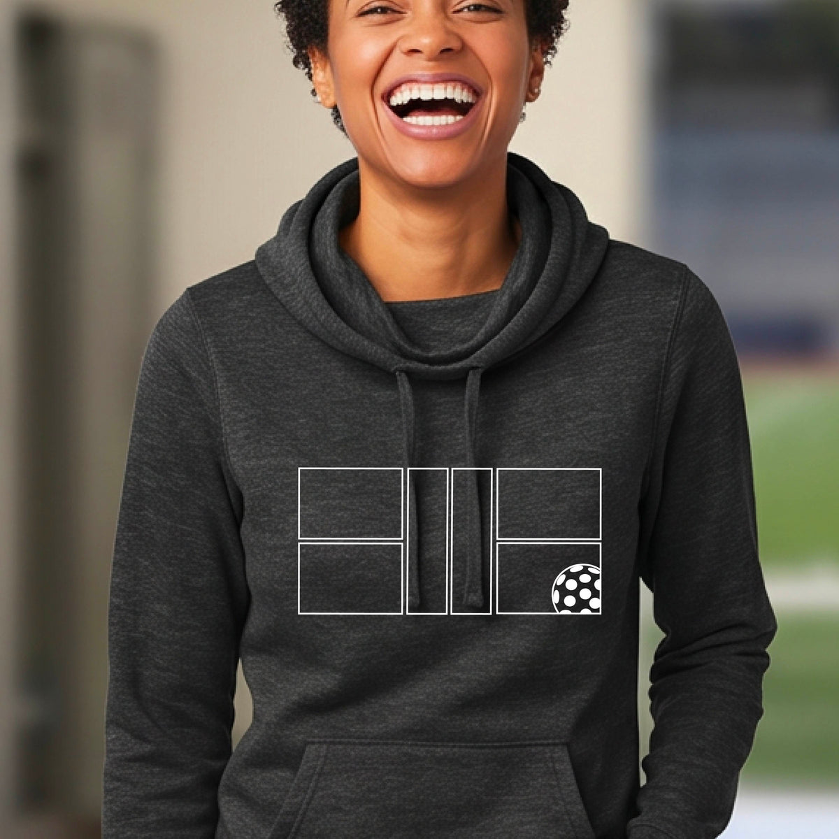 A woman wearing a  heather black cowlneck hoodie with a white graphic print of a pickleball court and a  pickleball graphic on the front, set against a blurred outdoor background.