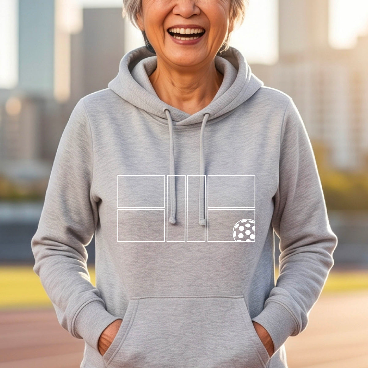A woman wearing a gray cowlneck hoodie with a white graphic print of a pickleball court and a  pickleball graphic on the front, set against a blurred outdoor background.