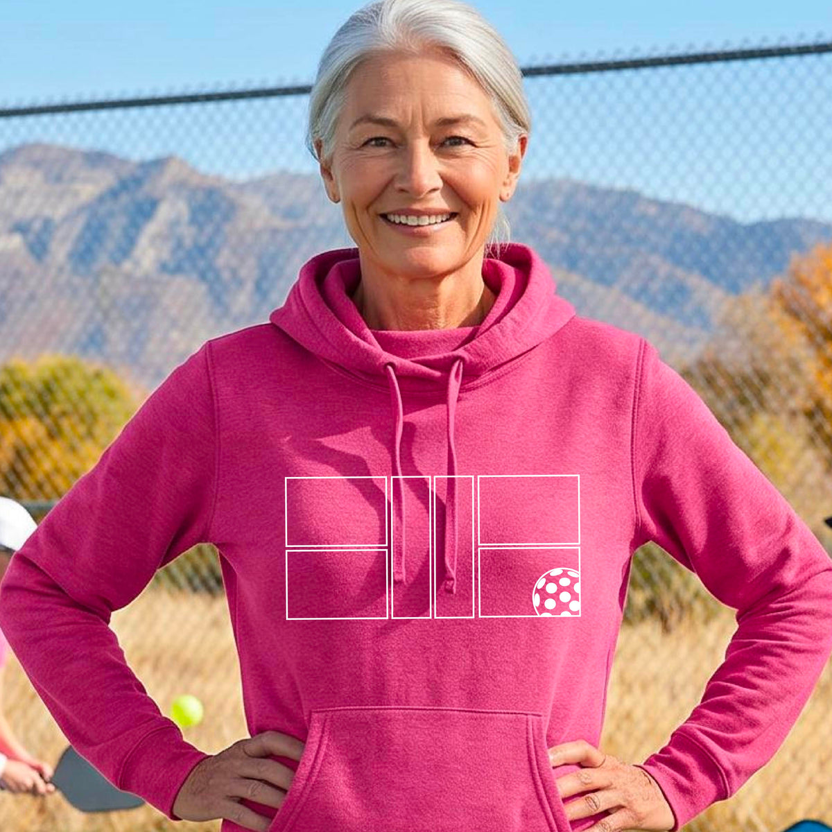 A woman wearing a pink cowlneck hoodie with a white graphic print of a pickleball court and a  pickleball graphic on the front, set against a blurred outdoor background.