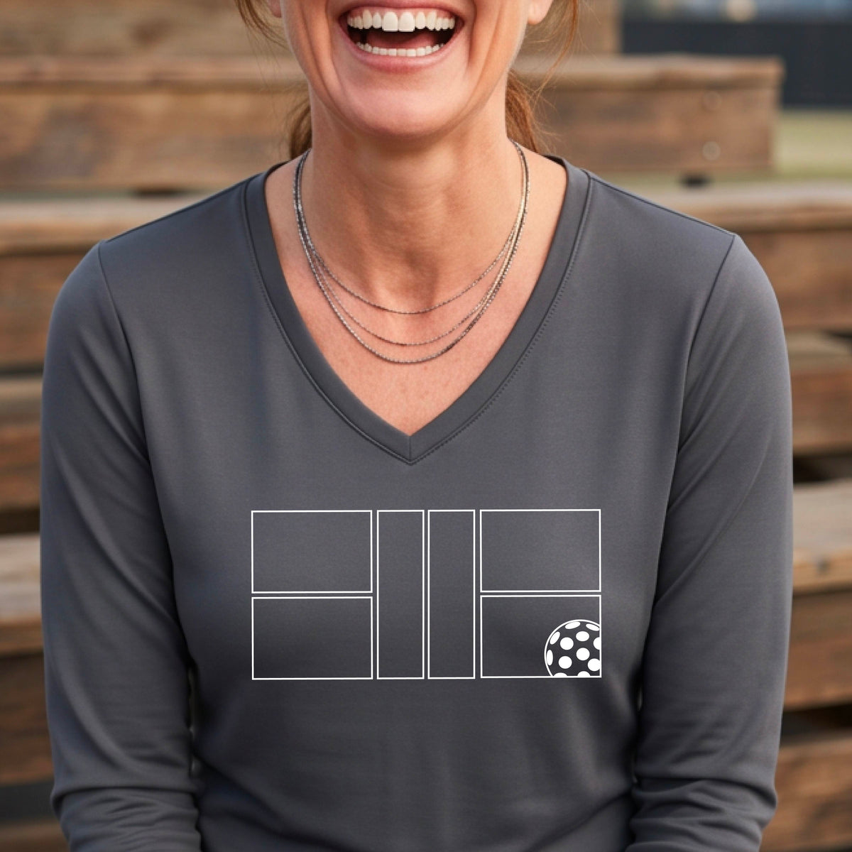 A woman on a pickleball court wearing a gray long-sleeve V-neck athletic shirt with a white pickleball court line graphic and a pickleball detail on the front.