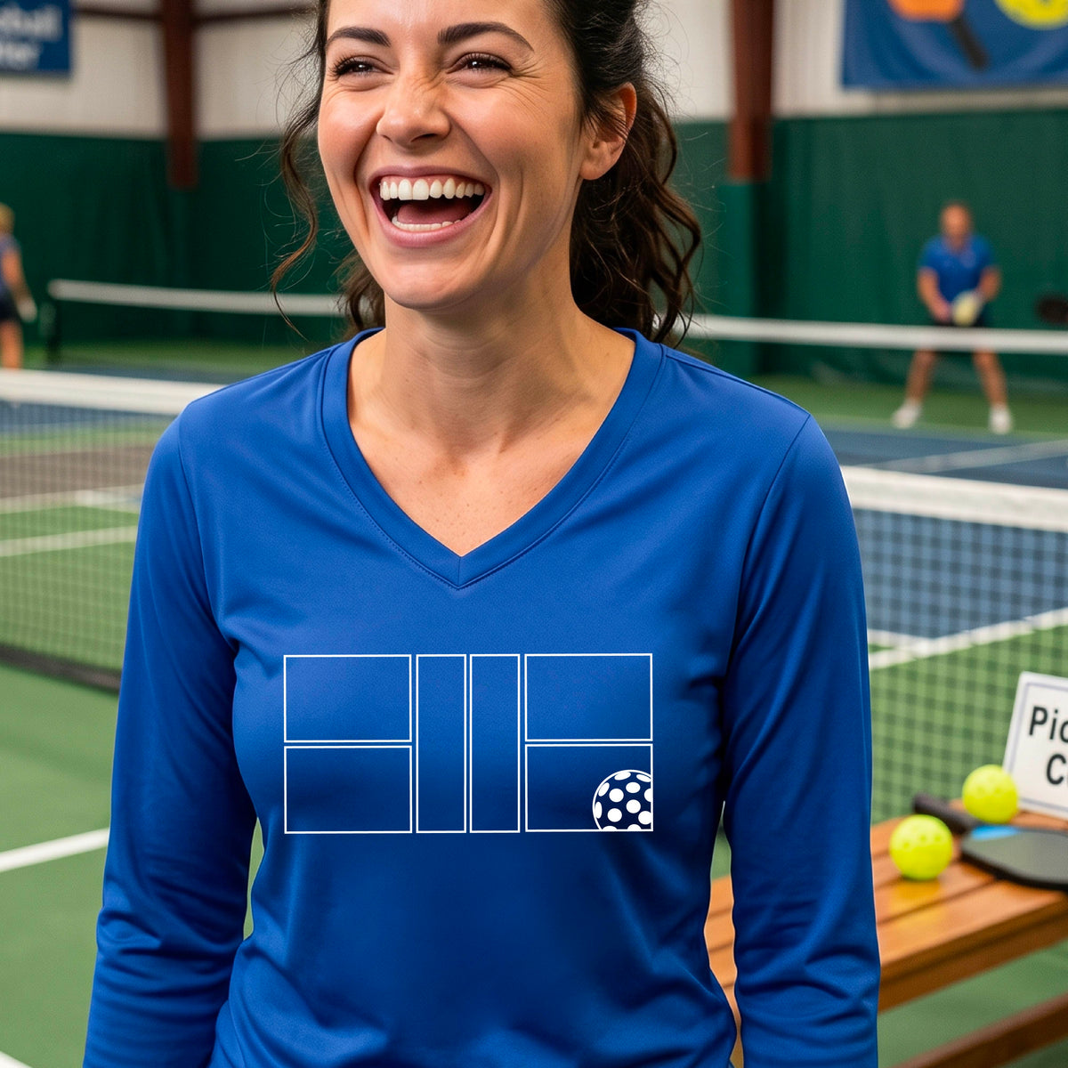 A woman on a pickleball court wearing a royal blue long-sleeve V-neck athletic shirt with a white pickleball court line graphic and a pickleball detail on the front.