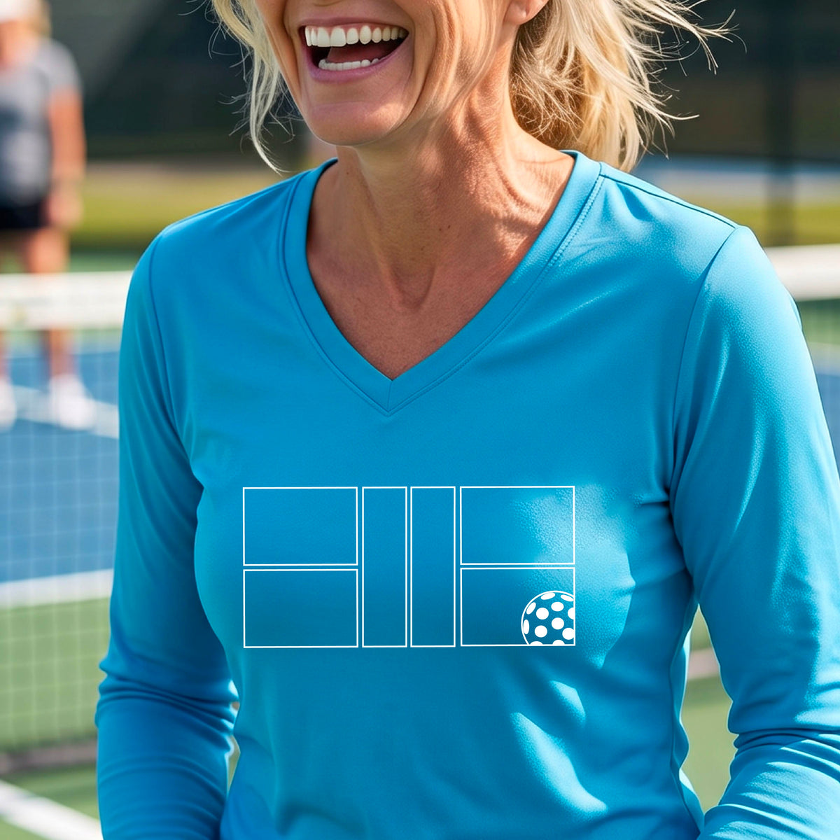 A woman on a pickleball court wearing a blue long-sleeve V-neck athletic shirt with a white pickleball court line graphic and a pickleball detail on the front.