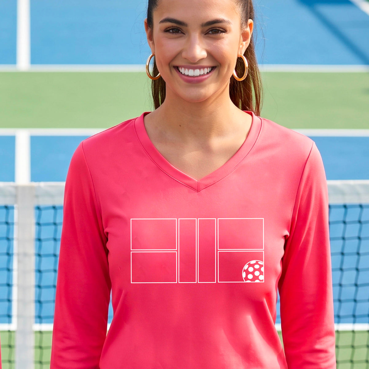 A woman on a pickleball court wearing a coral pink long-sleeve V-neck athletic shirt with a white pickleball court line graphic and a pickleball detail on the front.