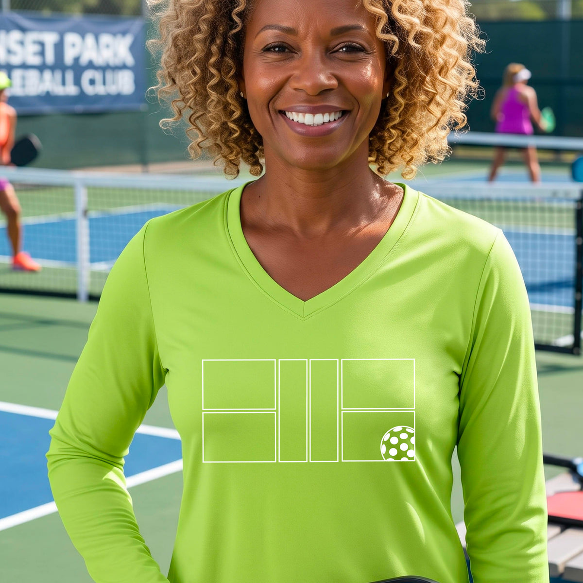 A woman on a pickleball court wearing a lime green long-sleeve V-neck athletic shirt with a white pickleball court line graphic and a pickleball detail on the front.