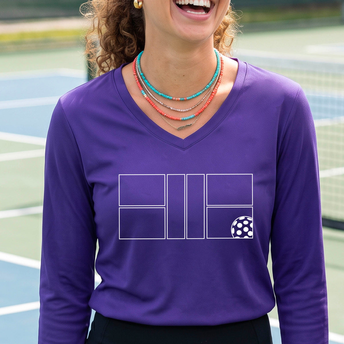 A woman on a pickleball court wearing a purple long-sleeve V-neck athletic shirt with a white pickleball court line graphic and a pickleball detail on the front.