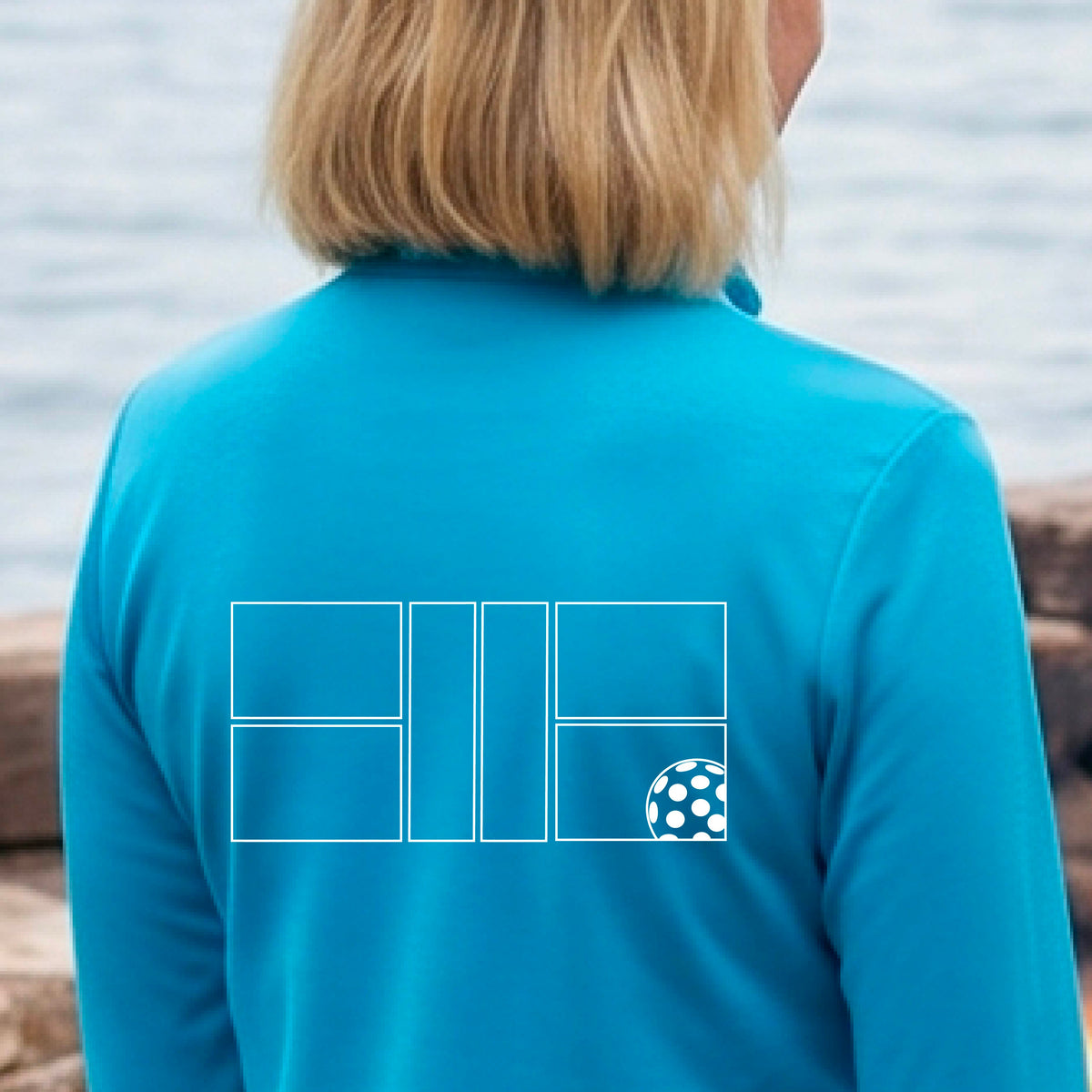 A woman on a pickleball court wearing a bright blue long-sleeve athletic quarter-zip jacket with a white geometric line pickleall court and ball graphic on the back.