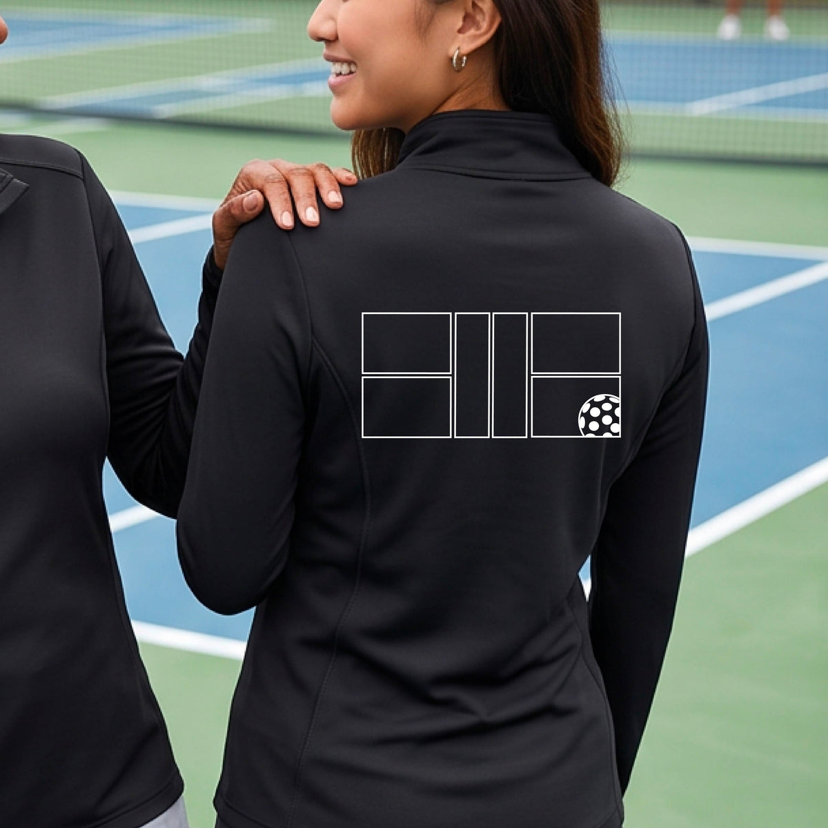 A woman on a pickleball court wearing a black long-sleeve athletic quarter-zip jacket with a white geometric line pickleall court and ball graphic on the back.