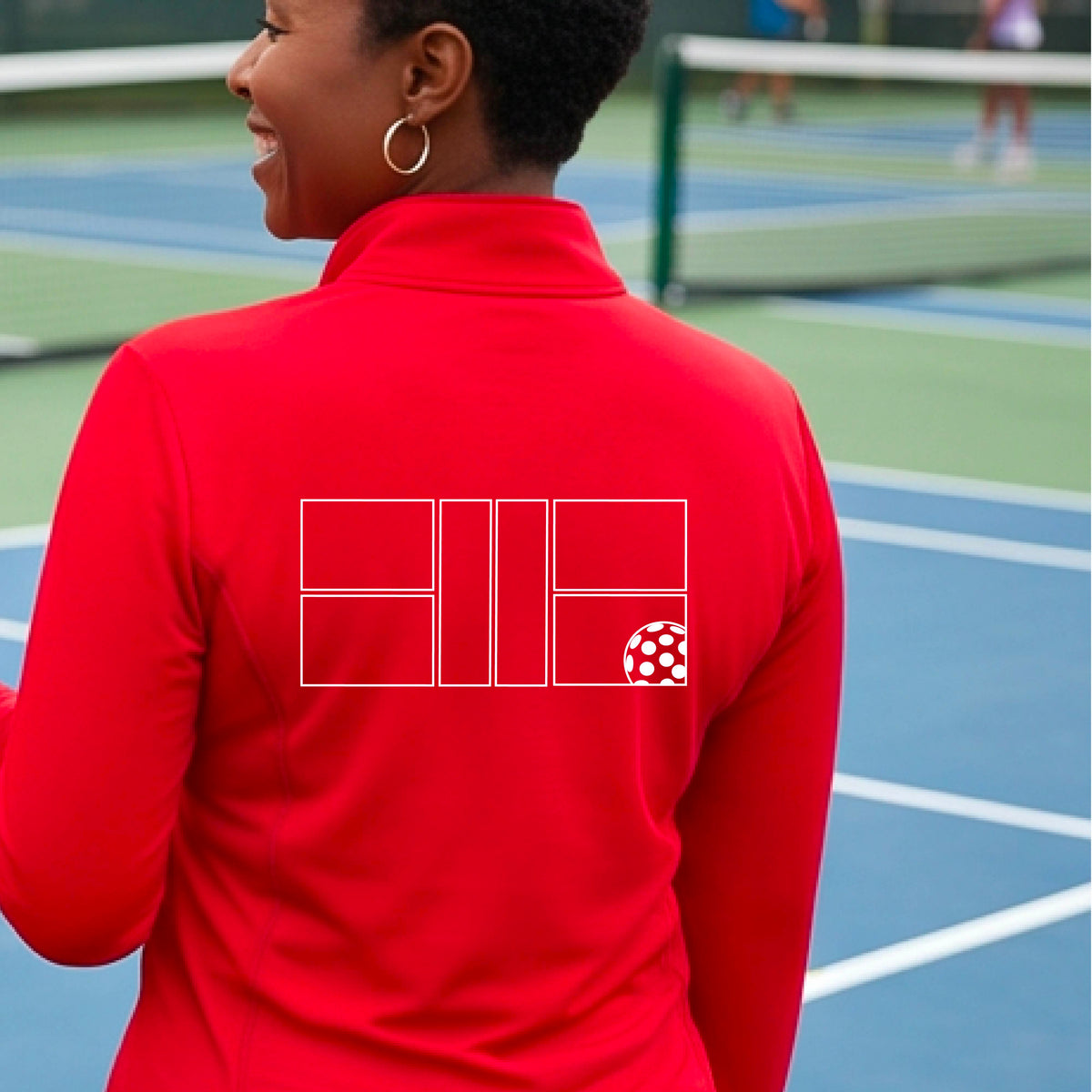 A woman on a pickleball court wearing a red long-sleeve athletic quarter-zip jacket with a white geometric line pickleall court and ball graphic on the back.