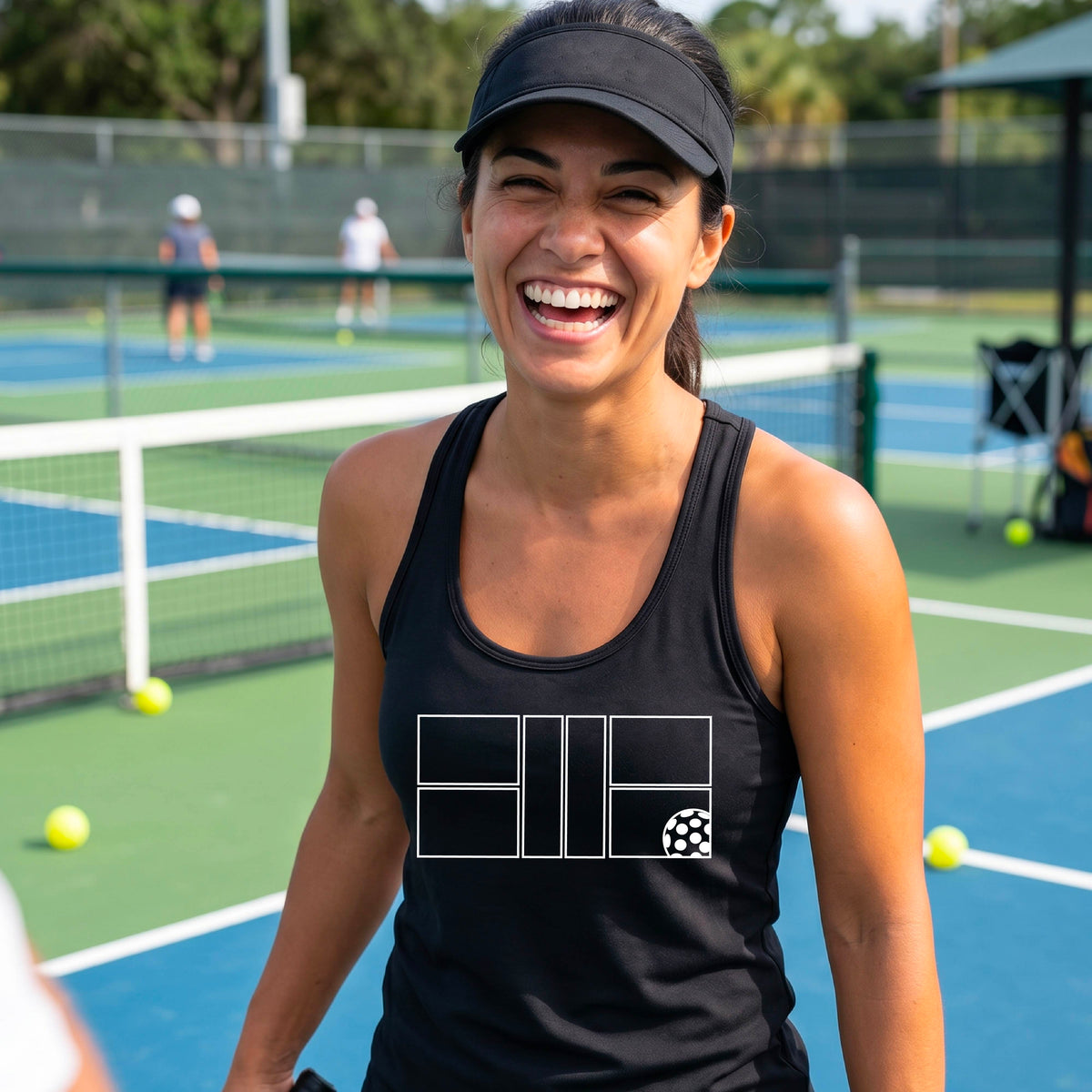 A smiling woman on a pickelball court wearing a black athletic tank top with a white graphic logo of a pickleball court and ball in front of a pickleball court. 