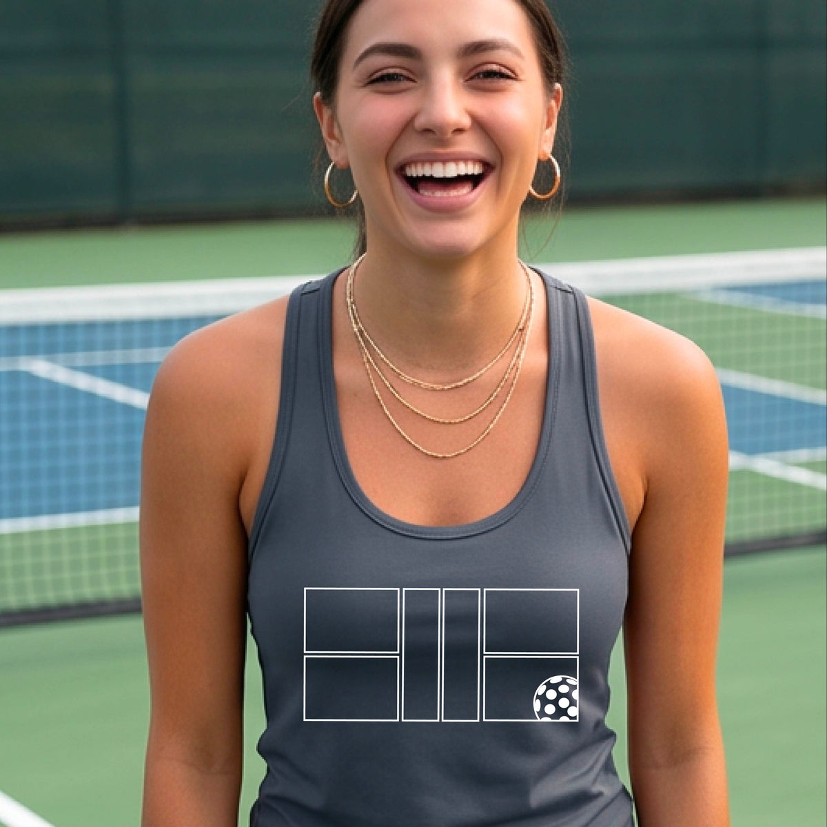 A smiling woman on a pickelball court wearing a gray athletic tank top with a white graphic logo of a pickleball court and ball in front of a pickleball court. 