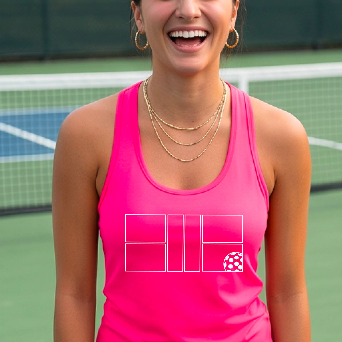 A smiling woman on a pickelball court wearing a pink athletic tank top with a white graphic logo of a pickleball court and ball in front of a pickleball court. 