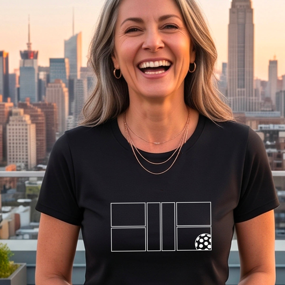 A smiling woman wearing a black crewneck t-shirt with a white graphic print on the front of a pwhite ickleball court and ball on the front. ,