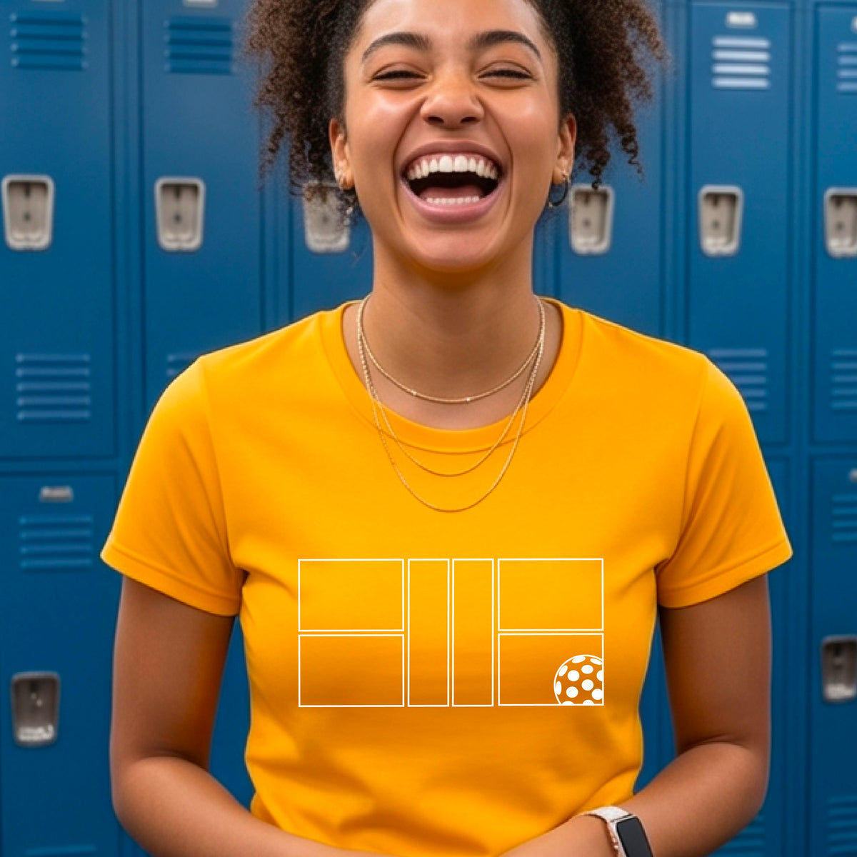 A smiling woman wearing a mustard yellow gold crewneck t-shirt with a white graphic print on the front of a pwhite ickleball court and ball on the front.
