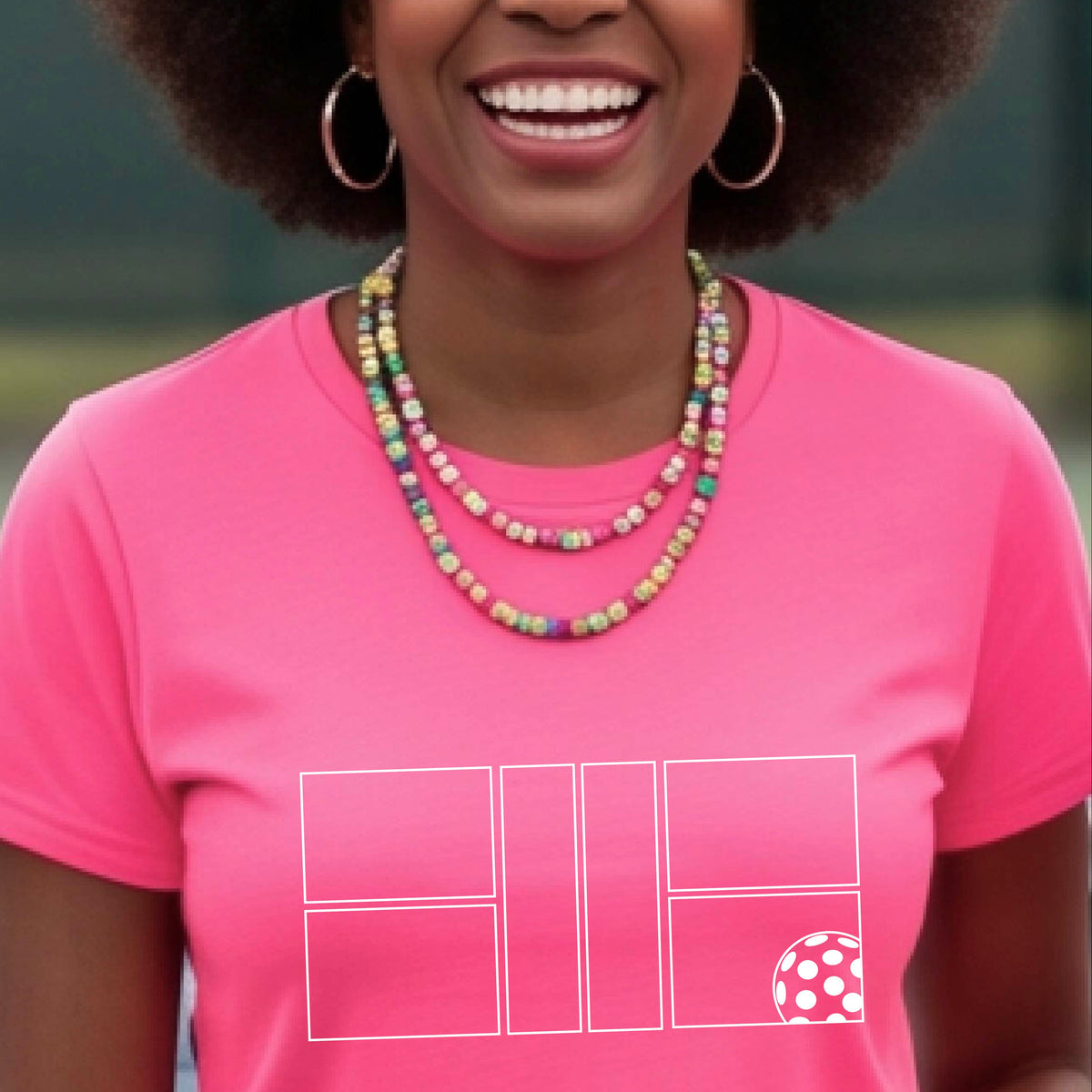 A smiling woman wearing a bright pink crewneck t-shirt with a white graphic print on the front of a pwhite ickleball court and ball on the front. ,