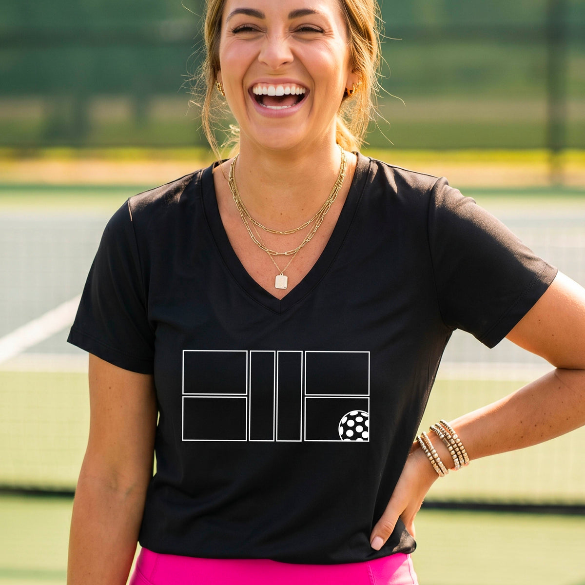 A smiling  woman on a pickleball court wearing a black V-neck athletic t-shirt with a white geometric pickleball court and  ball graphic on the front.
