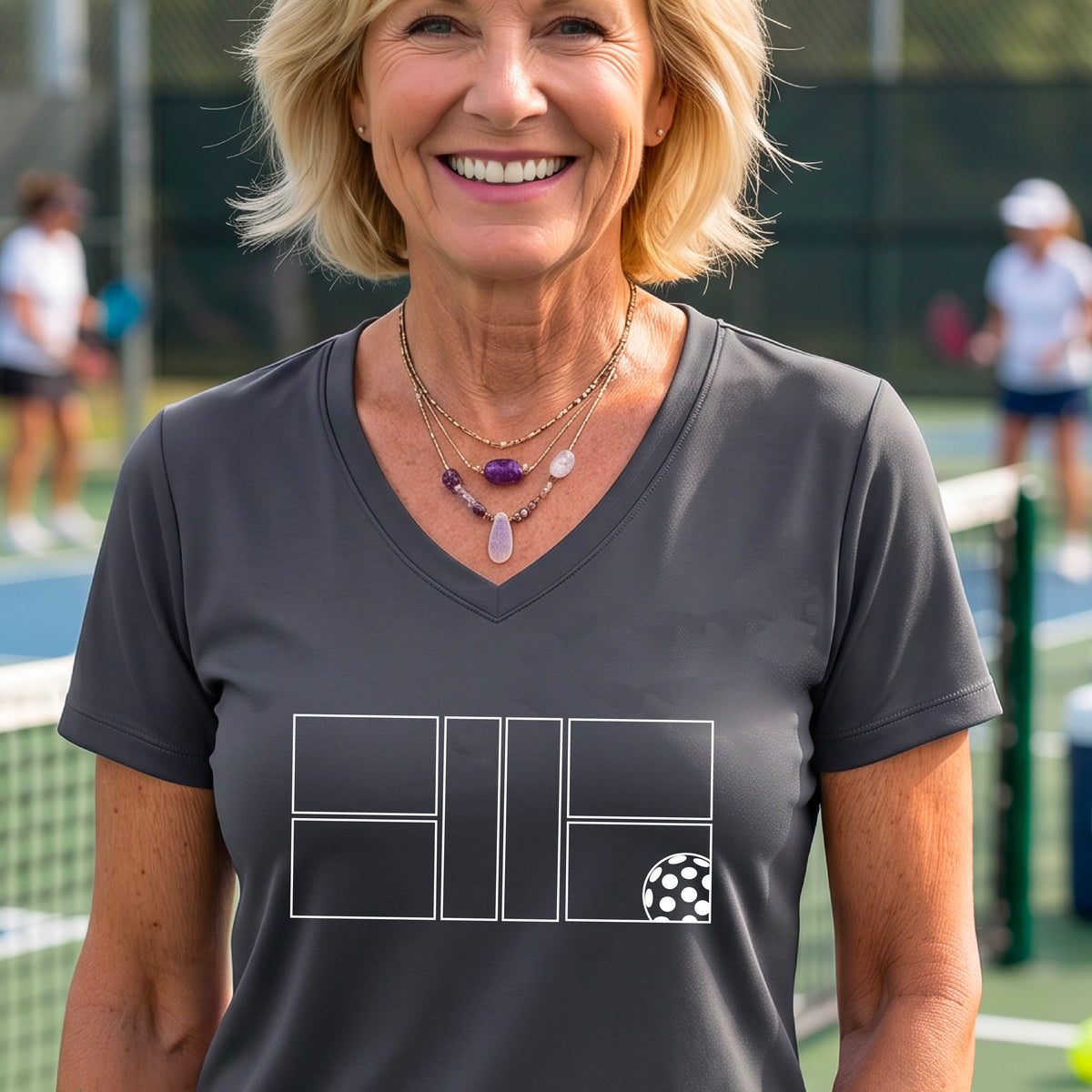 A smiling  woman on a pickleball court wearing a dark gray V-neck athletic t-shirt with a white geometric pickleball court and  ball graphic on the front.
