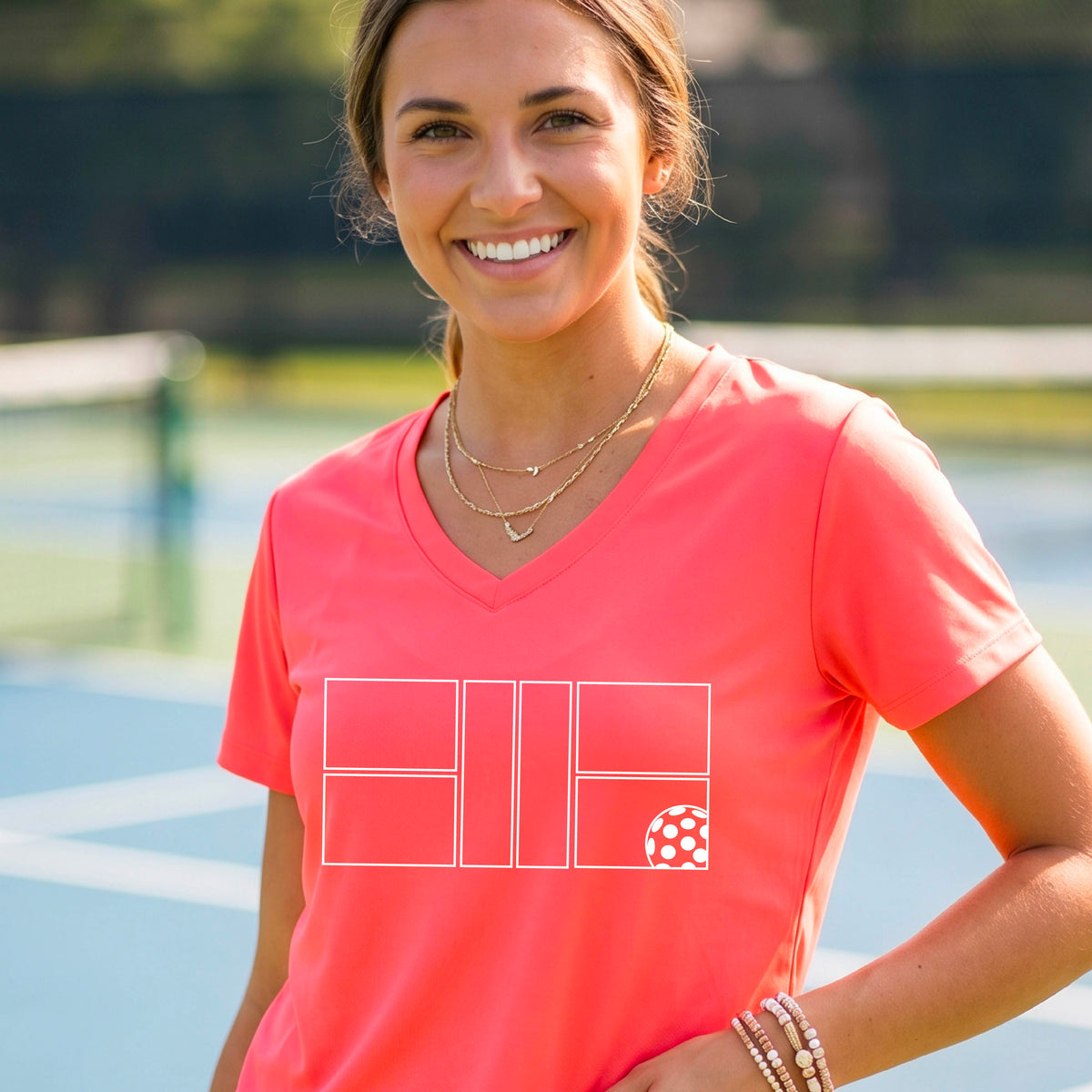 A smiling  woman on a pickleball court wearing a bright coral pink V-neck athletic t-shirt with a white geometric pickleball court and  ball graphic on the front.