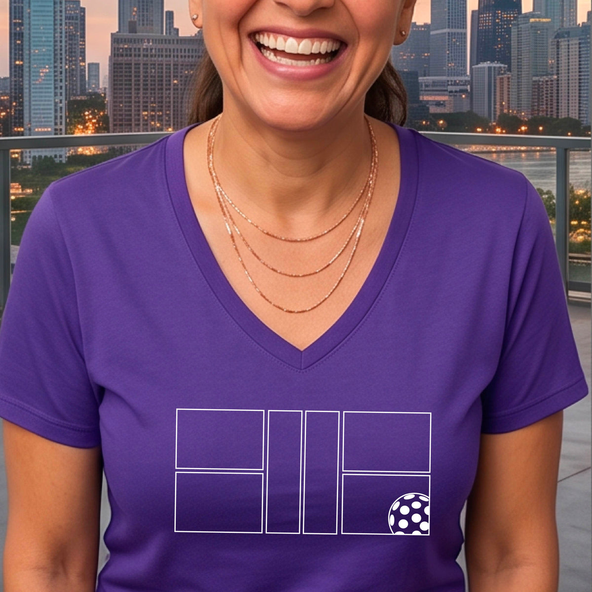 A smiling  woman on a pickleball court wearing a purple V-neck athletic t-shirt with a white geometric pickleball court and  ball graphic on the front.