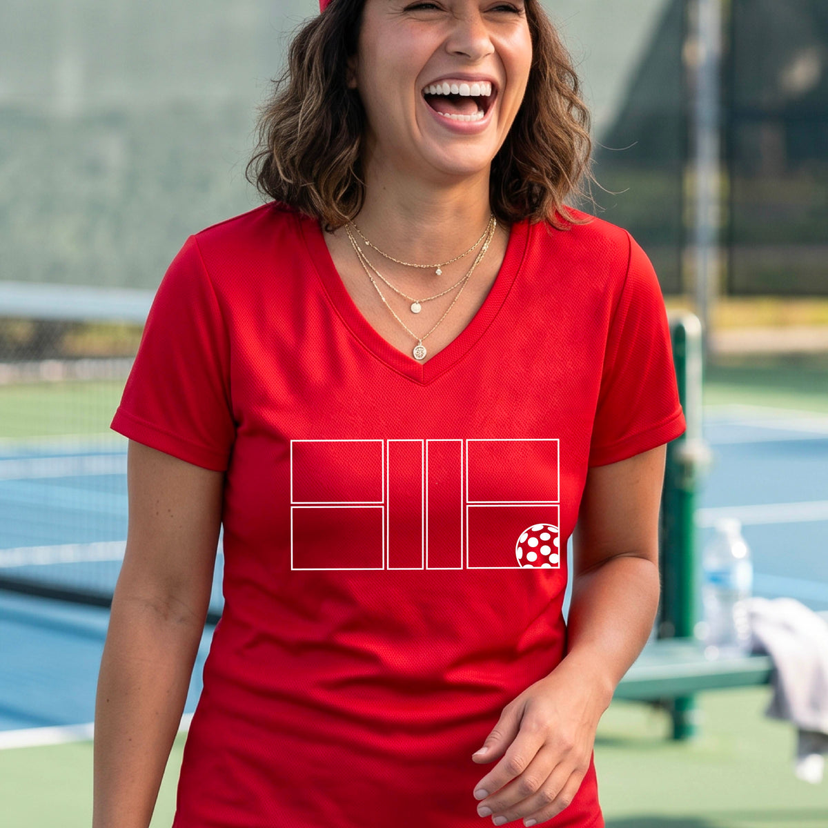 A smiling  woman on a pickleball court wearing a red V-neck athletic t-shirt with a white geometric pickleball court and  ball graphic on the front.