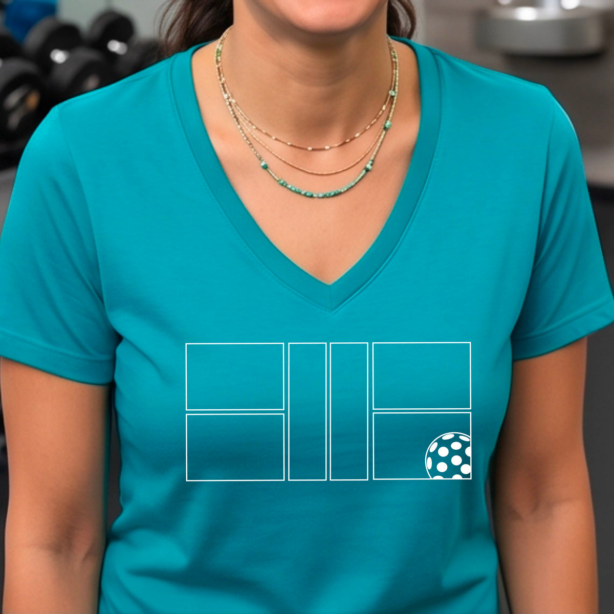 A smiling  woman on a pickleball court wearing a teal V-neck athletic t-shirt with a white geometric pickleball court and  ball graphic on the front.