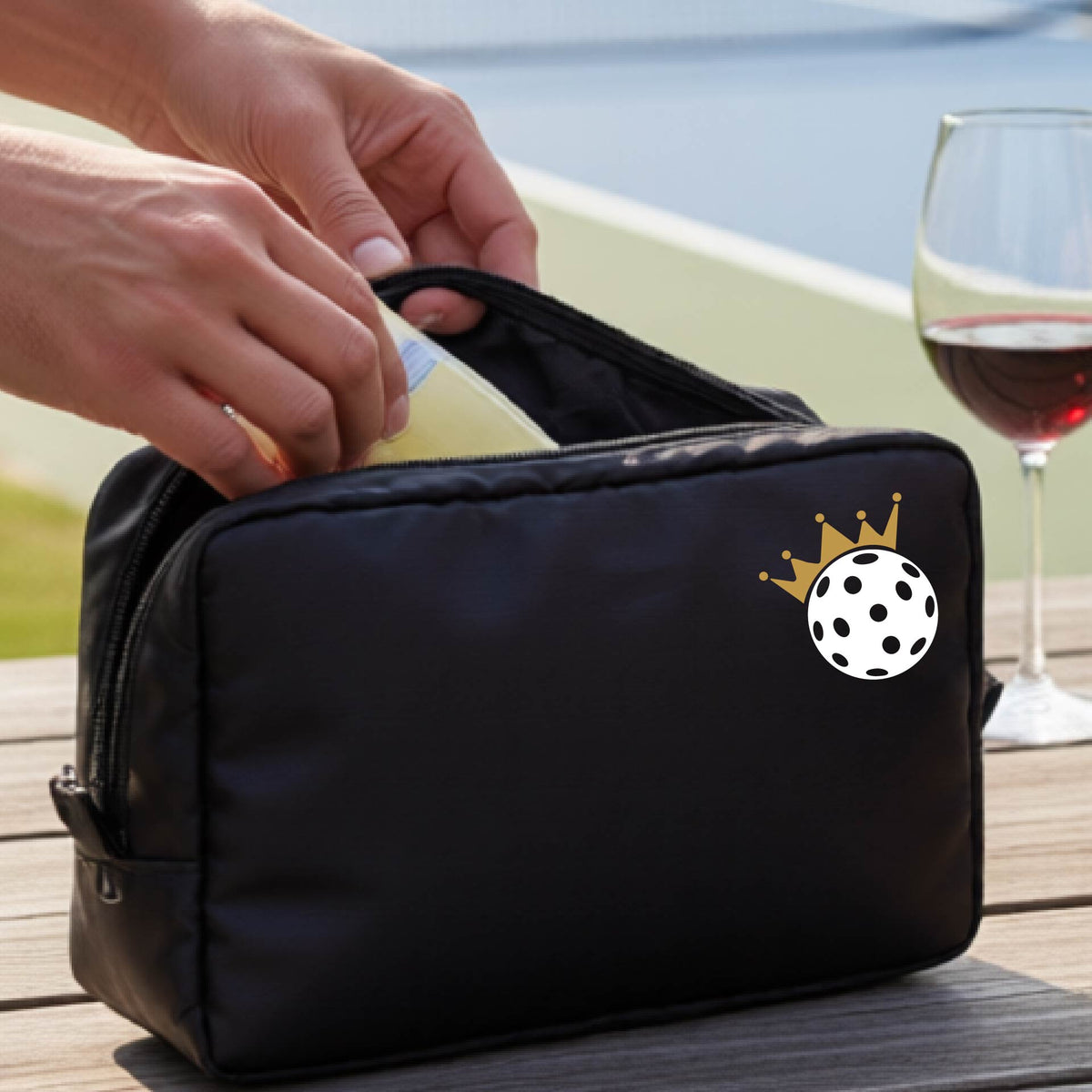 A person holds a black zippered pouch with a small white pickleball and gold crown graphic on the front, set against an outdoor background with a pickleball court and wooden table.