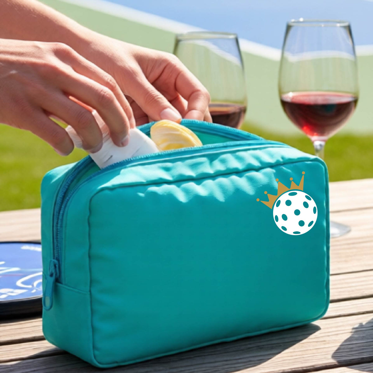 A person holds a turquoise zippered pouch with a small white pickleball and gold crown graphic on the front, set against an outdoor background with a pickleball court and wooden table.