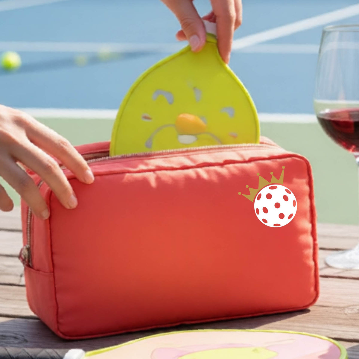 A person holds a coral colored zippered pouch with a small white pickleball and gold crown graphic on the front, set against an outdoor background with a pickleball court and wooden table.
